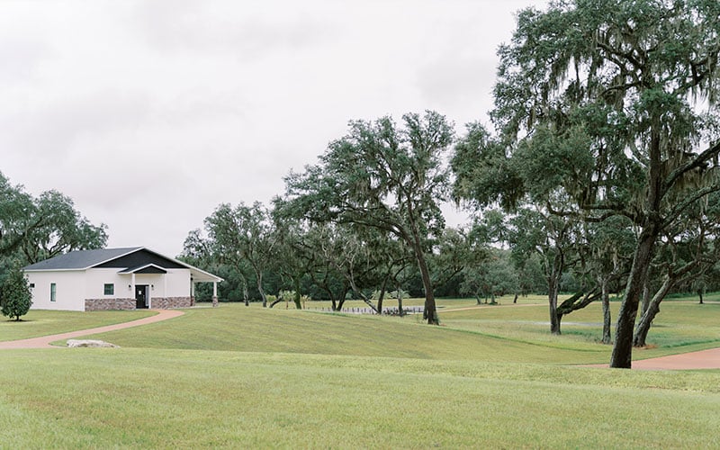 A brick and white house in a field of grass that is flanked with oak trees.